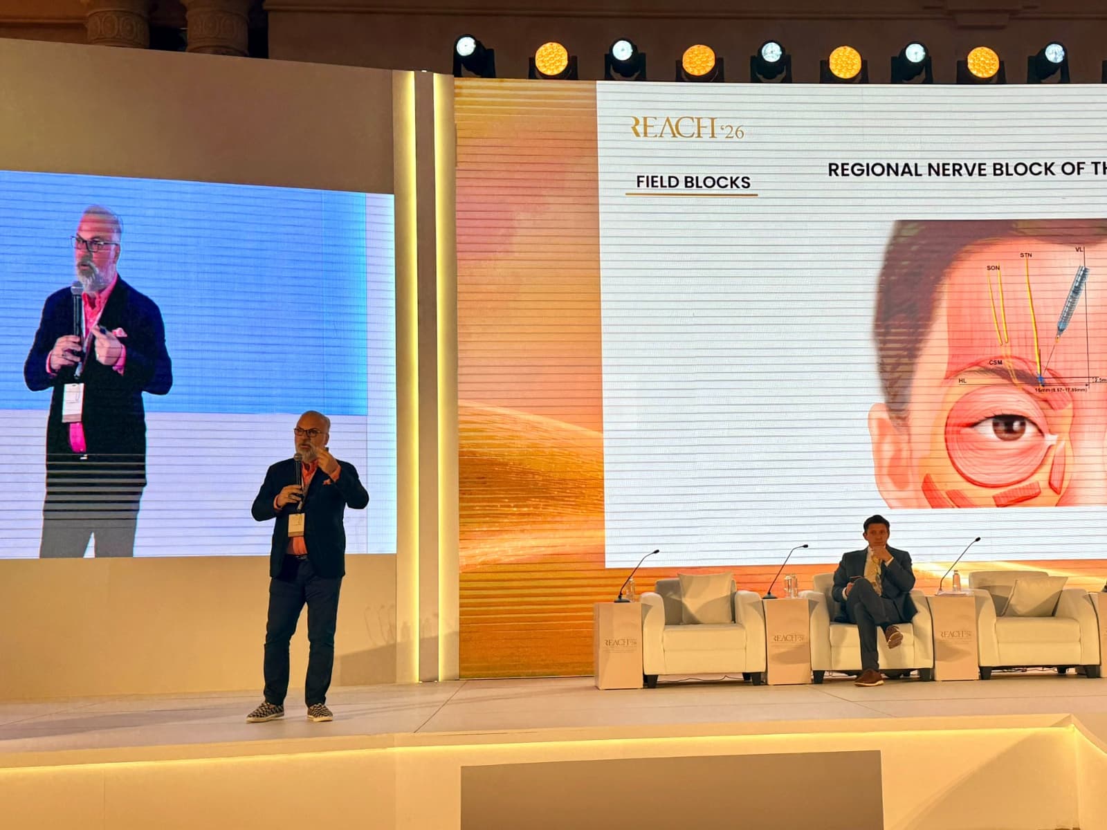 Speaker on stage at REACH 2026 presenting medical techniques for local anesthesia.